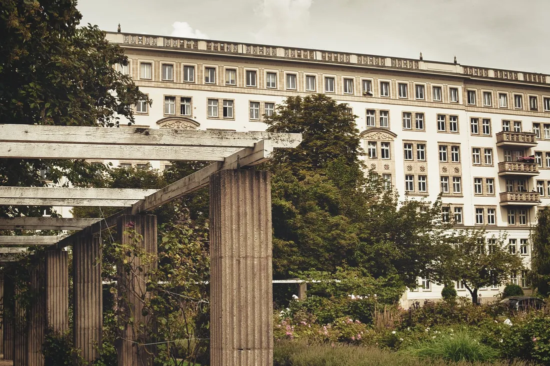 Langgestreckte Altbaufassade mit Balkonen hinter Bäumen und einer Pergola